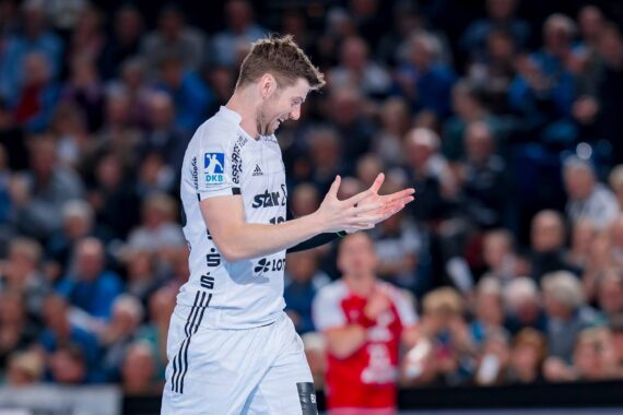 Ein männlicher Handballspieler in einem weißen Trikot klatscht während eines Spiels in die Hände und lächelt, während im Hintergrund eine verschwommene Menschenmenge und ein weiterer Spieler in Rot zu sehen sind.