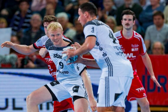 Vier männliche Handballspieler, zwei in weißen und zwei in roten Trikots, befinden sich während eines Spiels im aktiven Wettkampf. Die Szene ist intensiv, und im unscharfen Hintergrund sind Zuschauer zu sehen.