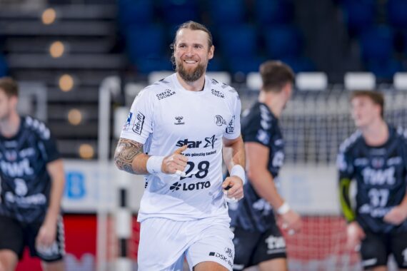 Ein männlicher Handballspieler in einem weißen Trikot mit der Nummer 28 lächelt, während er auf dem Spielfeld läuft. Im Hintergrund sind unscharfe Mitspieler und Gegner in dunklen Trikots und ein Tor zu sehen.