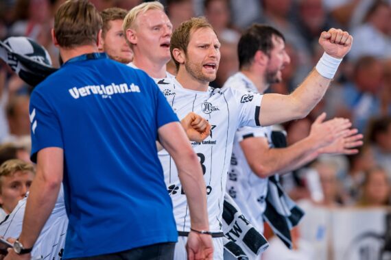 Eine Gruppe von männlichen Handballspielern in weißen Trikots jubelt während eines Spiels am Spielfeldrand, wobei ein Spieler vorne die Faust zum Jubeln hebt. Im Hintergrund ist die Zuschauermenge zu sehen.