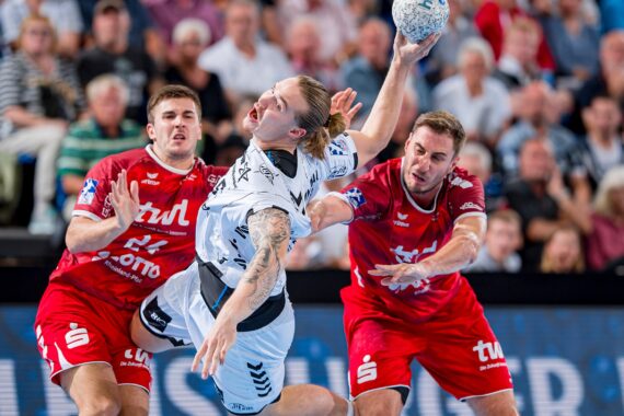 Ein Handballspieler in einem weißen Trikot springt mit dem Ball, umgeben von zwei Verteidigern in roten Trikots, während eines intensiven Spiels. Im Hintergrund sieht eine Gruppe von Zuschauern zu.