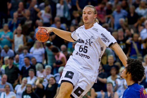 Ein männlicher Handballspieler in einem weißen Trikot mit der Nummer 65 bereitet sich darauf vor, den Ball während eines Spiels zu werfen, während ein Verteidiger in einem blauen Trikot versucht, ihn zu blockieren. Die Zuschauer füllen die Tribünen im Hintergrund.