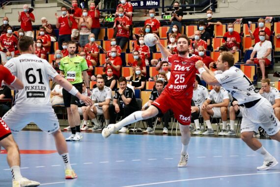 Ein Handballspieler in einem roten Trikot springt und bereitet sich darauf vor, den Ball zu werfen, während er von Spielern in weißen Trikots verteidigt wird; im Hintergrund sind ein Schiedsrichter, Mannschaftsbänke und maskierte Zuschauer zu sehen.