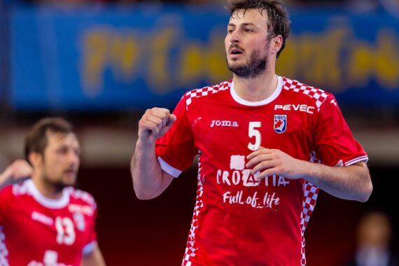 Ein männlicher Handballspieler, der ein rotes Kroatien-Trikot mit der Nummer 5 trägt, steht im Mittelpunkt und wirkt während eines Spiels entschlossen. Ein anderer Spieler in einem ähnlichen Trikot ist im Hintergrund zu sehen.