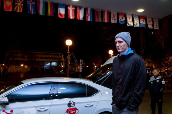 Ein Mann mit grauer Mütze und schwarzer Jacke steht nachts in der Nähe eines weißen Autos, mit einer weiteren Person im Hintergrund. Über ihnen hängen verschiedene Nationalflaggen.
