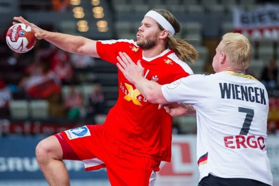 Zwei männliche Handballspieler in Aktion: Der eine in einem roten Trikot springt und bereitet sich auf einen Schuss vor, während ein Spieler in einem weißen Trikot mit der Aufschrift WIENCEK 7 ihn verteidigt. Im Hintergrund ist eine Sporthalle zu sehen.