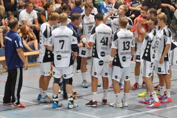 Eine Herren-Handballmannschaft in weißen Trikots kauert auf einem Hallenplatz zusammen und hört einem Trainer in einem blauen Hemd zu. Die Zuschauer schauen von der Tribüne im Hintergrund zu.