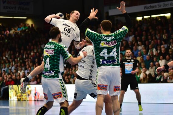 Ein Handballspieler in einem weißen Trikot springt zum Schuss, während drei Verteidiger in grünen Trikots versuchen, ihn während eines Spiels in einer überfüllten Halle zu blockieren.