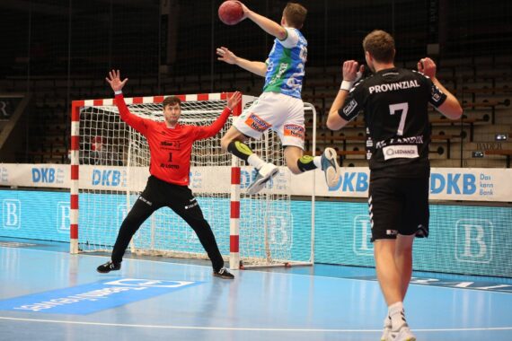 Ein Handballspieler in einem blau-weißen Trikot springt auf, um auf das Tor zu schießen, während der Torwart in Rot sich darauf vorbereitet, das Tor zu blockieren. Ein anderer Spieler in einem schwarzen Trikot steht in der Nähe auf dem Spielfeld.