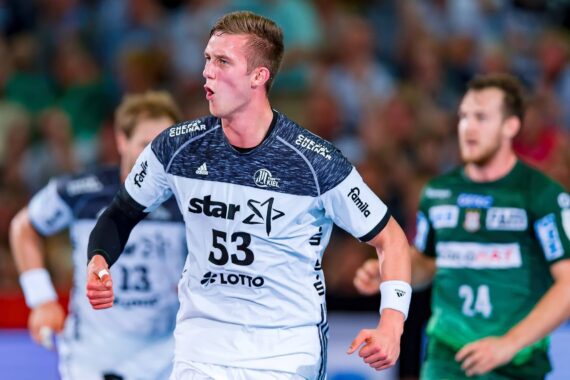 Ein männlicher Handballspieler in einem weiß-schwarzen Trikot mit der Nummer 53 jubelt auf dem Spielfeld, während zwei gegnerische Spieler in grünen Trikots und eine unscharfe Menschenmenge im Hintergrund zu sehen sind.