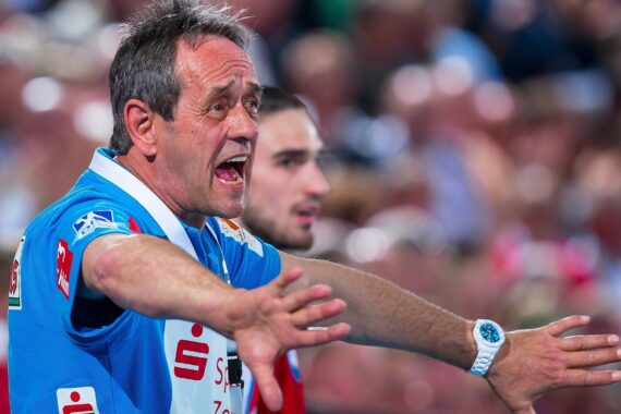 Ein Mann in einem blauen Sporttrikot gestikuliert mit beiden Armen und scheint während einer Sportveranstaltung zu rufen oder Anweisungen zu geben. Der Hintergrund ist mit Zuschauern unscharf.