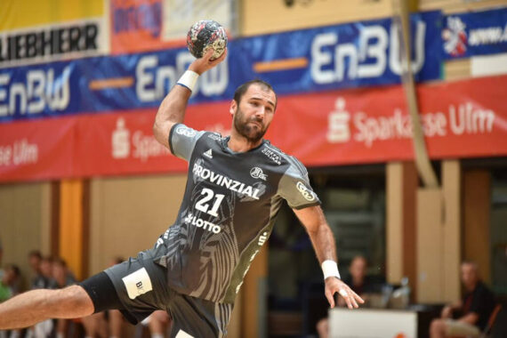Ein männlicher Handballspieler in einem grauen Trikot mit der Nummer 21 befindet sich mit erhobenem Arm in der Luft und bereitet sich auf einen Wurf während eines Spiels vor. Im Hintergrund sind die Halle und die Zuschauer zu sehen.