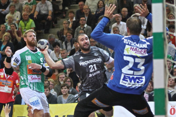 Ein Handballspieler in einem schwarzen Trikot springt, um den Ball zu schießen, während ein Verteidiger in Grün und ein Torwart in Blau versuchen, ihn zu blockieren; ein Zuschauer beobachtet das Geschehen im Hintergrund.