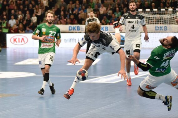 Ein Handballspieler in einem weiß-schwarzen Trikot springt nach vorne, um den Ball zu werfen, während ein Verteidiger in einem grünen Trikot versucht, ihn zu blockieren. Andere Spieler und Zuschauer sind im Hintergrund auf dem Spielfeld zu sehen.