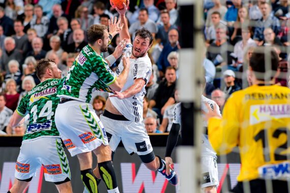 Eine Gruppe von Handballspielern in Aktion. Ein Spieler in Weiß springt, um den Ball zu schießen, während zwei Spieler in Grün verteidigen. Ein Torwart in Gelb steht vor dem Netz bereit. Die Zuschauer im Hintergrund schauen aufmerksam zu.