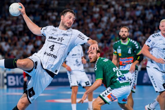 Ein Handballspieler in einem weißen Trikot springt in die Luft, um den Ball zu werfen, während Spieler in grünen und weißen Trikots während eines Spiels in einer überfüllten Halle zusehen.