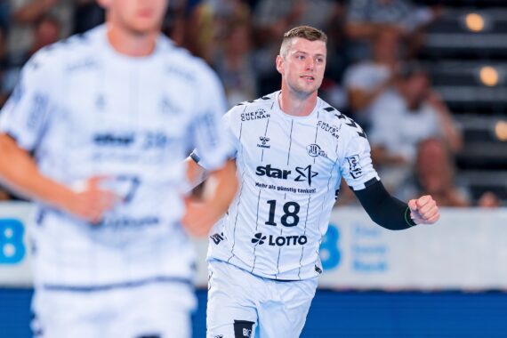 Ein männlicher Handballspieler in einem weiß-schwarz gestreiften Trikot mit der Nummer 18 läuft auf einem Hallenplatz, mit unscharfen Mitspielern und Zuschauern im Hintergrund.