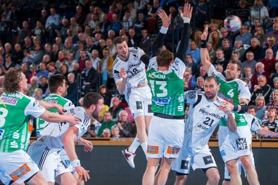 Eine Gruppe von Handballspielern in weiß-grünen Trikots liefert sich einen intensiven Wettkampf. Ein Spieler springt, um den Ball zu werfen, während die Zuschauer von den vollbesetzten Tribünen im Hintergrund zusehen.