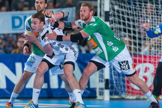 Drei männliche Handballspieler sind auf einem Hallenplatz in Aktion. Zwei Spieler in grünen Trikots verteidigen gegen einen Spieler in einem weißen Trikot, der den Ball hält. Die Szene ist in der Nähe des Torraums sehr intensiv.