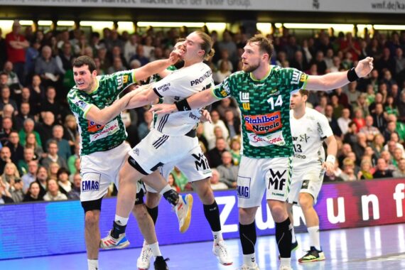 Drei männliche Handballspieler in Aktion während eines Spiels; ein Spieler in Weiß wird von zwei Spielern in grünen Trikots geblockt, wobei eine Hand das Gesicht des weißen Spielers berührt. Die Zuschauer füllen die Tribünen im Hintergrund.