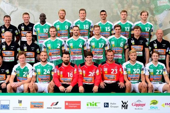Eine Handballmannschaft der Männer stellt sich für ein Gruppenfoto auf. Die Spieler in grün-weißen Trikots, zwei in roten Trikots und die Trainer in schwarzer Kleidung stehen und sitzen in drei Reihen. Die Logos der Sponsoren sind am unteren Rand zu sehen.