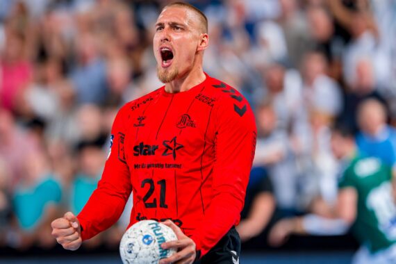 Ein männlicher Handballspieler in einem roten Trikot mit der Nummer 21 schreit leidenschaftlich, während er einen Handball hält. Der Hintergrund zeigt eine unscharfe Menge und andere Spieler in Aktion.