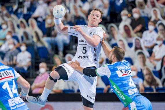 Ein Handballspieler in einem weißen Trikot springt und bereitet sich darauf vor, den Ball zu werfen, während zwei Spieler in blauen und grünen Trikots versuchen, ihn zu blockieren. Im Hintergrund ist ein Publikum zu sehen, von dem einige Masken tragen.