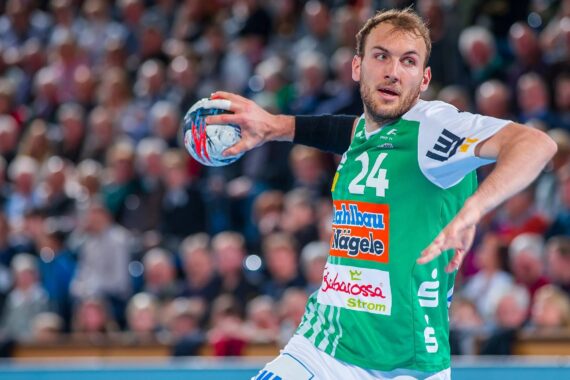 Ein männlicher Handballspieler in einem grün-weißen Trikot mit der Nummer 24 bereitet sich während eines Spiels auf den Wurf vor, während im Hintergrund eine unscharfe Menschenmenge zu sehen ist.
