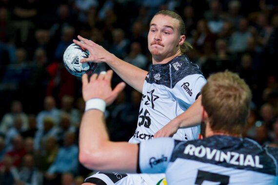 Ein Handballspieler in einem weiß-schwarzen Trikot bereitet sich darauf vor, den Ball zu werfen, während er von einem anderen Spieler in einem passenden Trikot während eines Spiels verteidigt wird, während die Zuschauer im Hintergrund zusehen.