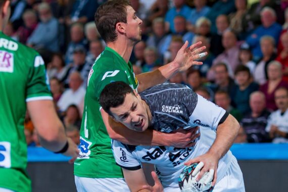Ein Handballspieler in einem weißen Trikot wird während eines Spiels von einem Gegner in einem grünen Trikot hart verteidigt, während im Hintergrund eine Zuschauermenge zusieht.