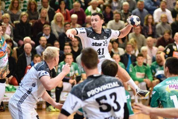 Ein Handballspieler in einem weißen Trikot springt, um den Ball zu werfen, während gegnerische Spieler in Grün und Mannschaftskameraden in Weiß zusehen. Die Zuschauer füllen die Tribünen im Hintergrund.