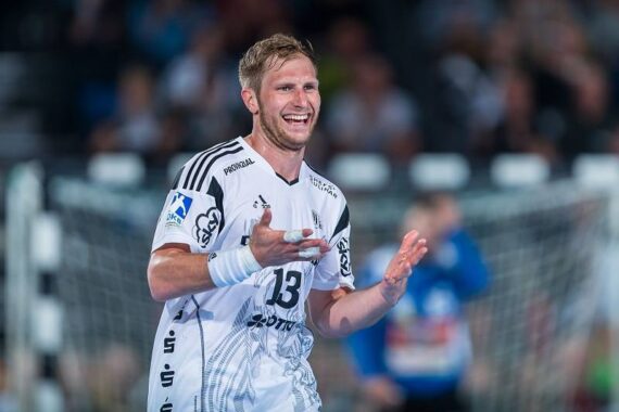 Ein männlicher Handballspieler in einem weißen Trikot mit der Nummer 13 lächelt und gestikuliert mit seinen Händen während eines Spiels, mit einem unscharfen Netz und Spielern im Hintergrund.