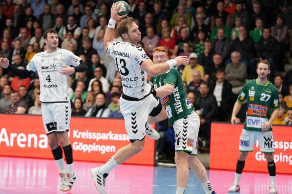 Ein Handballspieler in einem weißen Trikot springt zum Schuss, während ein Verteidiger in einem grünen Trikot versucht, den Ball zu blocken. Das Geschehen spielt sich auf einem Spielfeld ab, während im Hintergrund eine Zuschauermenge aufmerksam zusieht.