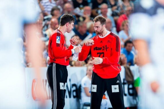 Zwei männliche Handballspieler in roten Trikots und schwarzen Hosen unterhalten sich während eines Spiels, wobei einer eine Wasserflasche in der Hand hält. Der Hintergrund zeigt eine unscharfe Menge und andere Spieler.