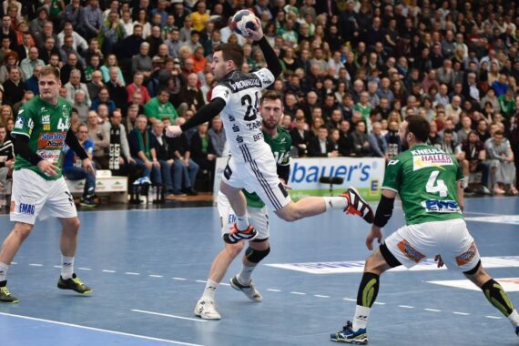 Ein Handballspieler in einem weißen Trikot springt, um den Ball zu schießen, während zwei Spieler in Grün versuchen, ihn zu blockieren. Das Spiel findet in einer Halle statt, während im Hintergrund eine große Menschenmenge zuschaut.