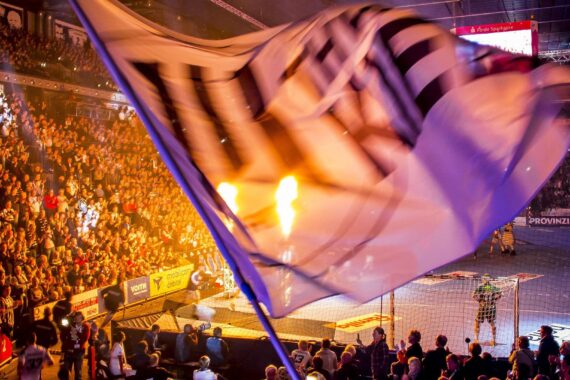 Eine überfüllte Sporthalle mit jubelnden Fans, einer großen wehenden Fahne im Vordergrund, hellen Lichtern und Flammen, die während einer lebhaften Veranstaltung in der Nähe des Torraums hochschlagen.