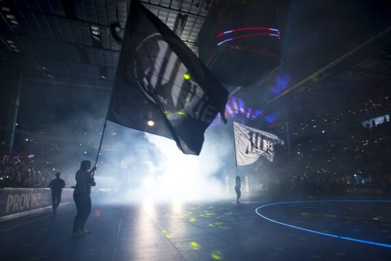 Menschen schwenken große Fahnen in einer schwach beleuchteten Sporthalle, umgeben von Rauch und dramatischer Beleuchtung, mit einer Menschenmenge und beleuchteten Bildschirmen im Hintergrund.