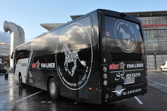 Ein schwarz-weißer Tourbus mit Fan-Club- und Zebra-Grafiken ist auf einem Parkplatz vor einem modernen Gebäude geparkt. Auf dem Bus sind Logos, eine Telefonnummer und ein deutsches Nummernschild angebracht.