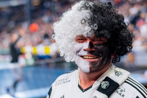 Eine lächelnde Person mit einer lockigen schwarz-weißen Perücke und passender Gesichtsbemalung trägt ein schwarz-weißes Sporttrikot und einen Schal und steht in einer überfüllten Arena oder einem Stadion.