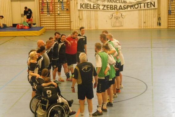 Eine Gruppe von Hallenhandballern, darunter ein Rollstuhlfahrer, steht im Kreis und legt die Arme umeinander. Hinter ihnen hängt ein Banner mit der Aufschrift Fanclub Kieler Zebrasprotten an der Wand.