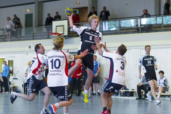 Ein Handballspieler in einem schwarz-weißen Trikot springt in der Luft, um den Ball zu werfen, während drei gegnerische Spieler in weißen Trikots versuchen, ihn während eines Hallenspiels zu blockieren. Zuschauer und Mannschaftskameraden schauen aus dem Hintergrund zu.