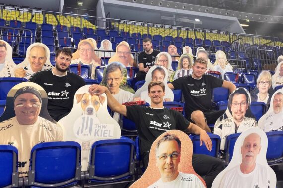 Vier Männer in schwarzen THW-Kiel-Trikots sitzen zwischen blauen Stadionsitzen, die mit großen Pappfiguren von Menschen und einem Hund gefüllt sind und den Anschein erwecken, dass das Publikum voll ist.