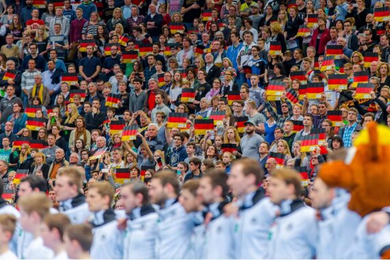 Eine große Menge von Fans in einem Stadion schwenkt enthusiastisch kleine deutsche Fahnen, gekleidet in Freizeitkleidung. Im Vordergrund stehen Sportler in weißen Uniformen und ein Maskottchen, die sich von der Menge abwenden.
