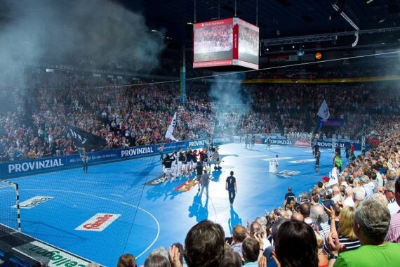 Eine große, mit Zuschauern gefüllte Halle, in der ein Handballspiel stattfindet. Auf dem blauen Spielfeld steigt Rauch auf, während sich Spieler und Offizielle versammeln, während über ihnen Banner und eine große Leinwand hängen.