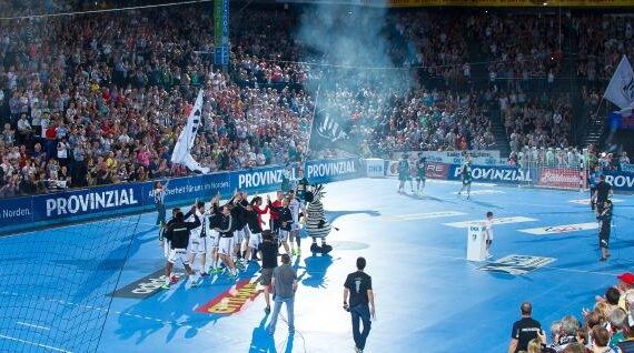 Eine große, mit Zuschauern gefüllte Halle umgibt ein blaues Handballfeld, in dessen Mitte sich Spieler und Offizielle zu einer Zeremonie versammeln. Helle Lichter und Fahnen sorgen für eine lebhafte Atmosphäre.