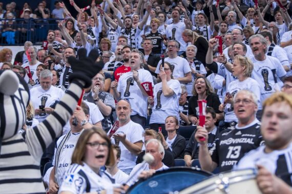 Eine große Menge begeisterter Sportfans, viele in weißen Hemden, jubeln und feiern in einem Stadion. Eine Person in einem Zebra-Maskottchen-Kostüm steht im Vordergrund in der Nähe einer Trommel und trägt zu der lebhaften Atmosphäre bei.