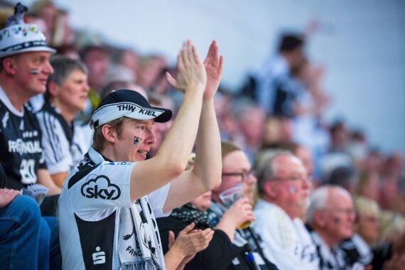 Eine Menge begeisterter Sportfans, viele in schwarz-weißer Mannschaftskleidung, klatschen und jubeln auf der Tribüne bei einer Hallenveranstaltung. Eine Person im Vordergrund trägt eine THW-Kiel-Mütze und ist geschminkt.
