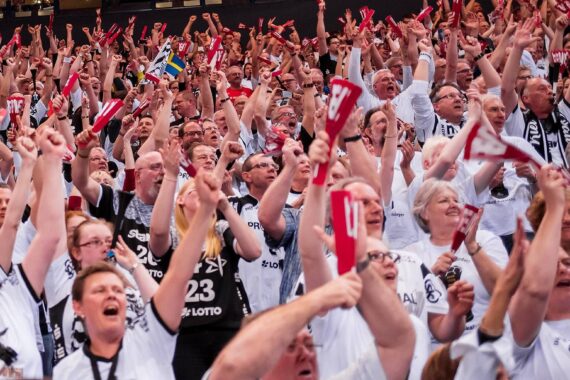 Eine große Menge begeisterter Sportfans in weißen Hemden jubelt und hebt rote Klatschen in einer Arena in die Luft, wodurch eine lebhafte und energiegeladene Atmosphäre entsteht.