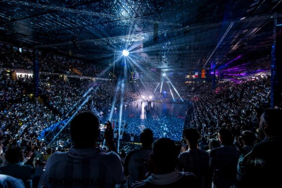 Ein überfülltes Hallenstadion mit jubelnden Fans und hellen Scheinwerfern, die die Bühne beleuchten und während einer lebhaften Veranstaltung oder eines Konzerts Lichtmuster auf der Menge und dem Boden erzeugen.