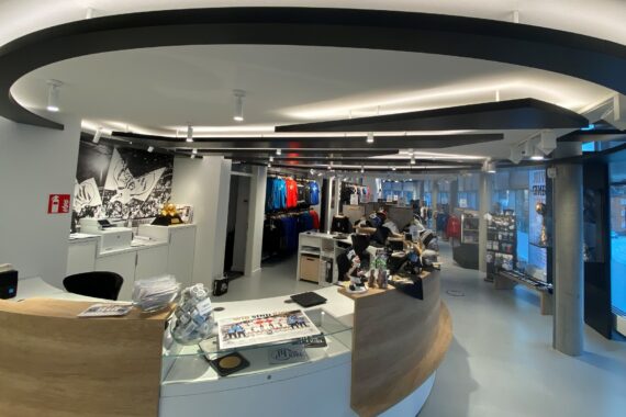 Moderne Einzelhandelseinrichtung mit geschwungenen Holztheken, Kleiderständern, Sportartikeln und einer Vitrine. Die Decke besteht aus schwarzen, gebogenen Balken mit integrierter Beleuchtung, die eine schlichte, offene Atmosphäre schaffen.